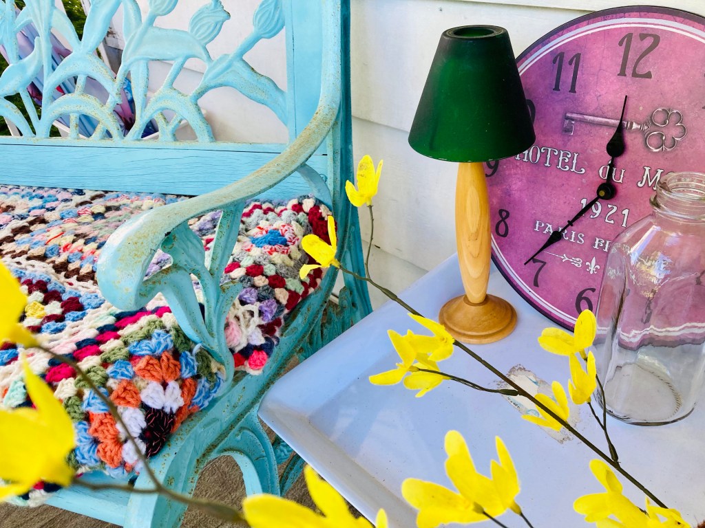 Part of a teal bench visible, with a small, white plastic table beside it. On the table are a candle lamp, clock, old milk jar, and some yellow flowers.