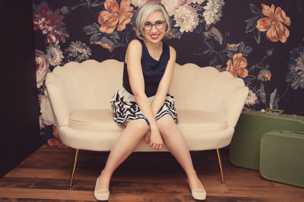 Portrait of Sonia Nicholson. She is seated on a bench with a scalloped back. The background is floral.