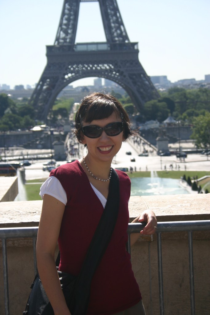 Woman in Paris, with Eiffel Tower in the background. She is wearing sunglasses and smiling.