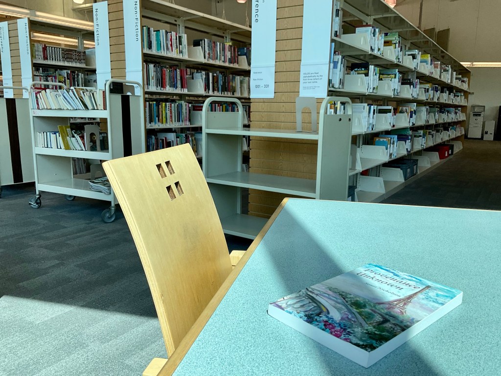 Copy of Provenance Unknown book by Sonia Nicholson on a table in a library. There are shelves of books in the background.