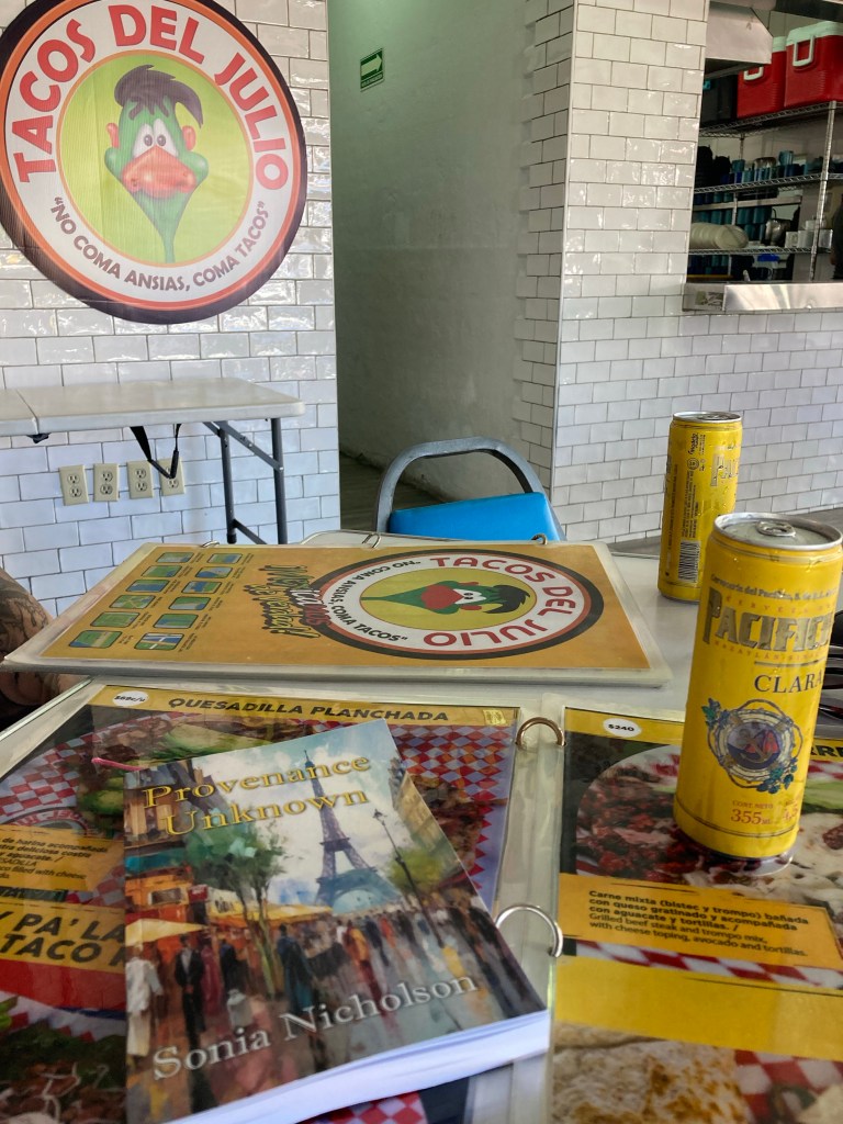 Copy of Provenance Unknown book by Sonia Nicholson on table of Tacos del Julio restaurant with menus and drinks.