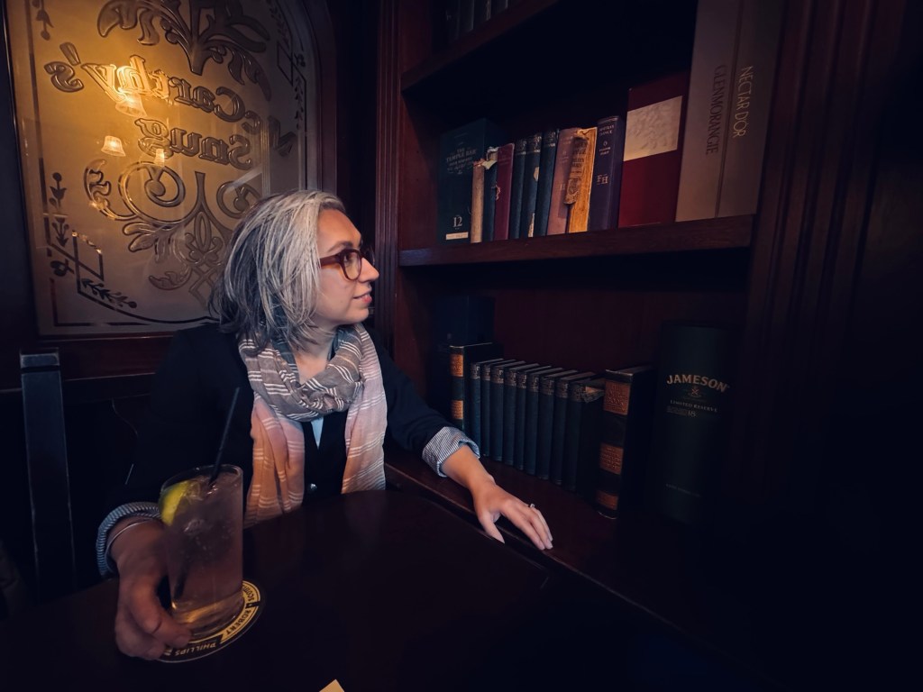 Author Sonia Nicholson sitting in dark Irish pub beside book shelves. 